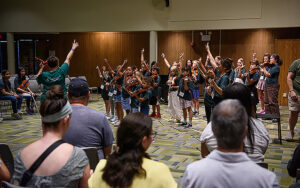 a group of young musicians with violins in the center of a large room while adult persons look on.